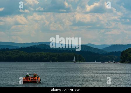 An einem schönen sonnigen Tag segeln die Menschen auf einem roten Tretboot mit Segelbooten und Booten im Hintergrund auf den See Stockfoto