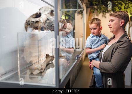 Neugieriger kleiner Junge in den Armen seiner Mutter, der ein Tierkelett in einer Knochenvitrine in einer Naturkundeausstellung ansieht. Stockfoto