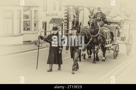 Begräbniscortège mit dem Begräbnisdirektor, der eine viktorianische Glaspferdewagen mit schwarzen Pferden und schwarzem Gefieder führt Stockfoto