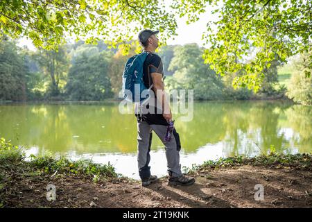 Seitenansicht des männlichen Walkers/ramblers mit Rucksack auf der Rückseite, mit Wasserbehälter, Blick auf einen britischen Süßwassersee an einem warmen, sonnigen Herbsttag. Stockfoto