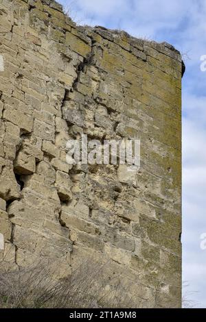 Eine alte moosbedeckte Steinmauer mit tiefen Rissen und eingestürzten Steinen auf dem Hintergrund eines bewölkten Himmels. Große diagonale Risse an der alten Wand. Spac kopieren Stockfoto