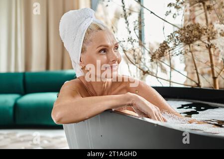 Eine Frau mittleren Alters mit weißem Handtuch auf dem Kopf, die in einem modernen Appartement ein Bad nimmt, Entspannung Stockfoto