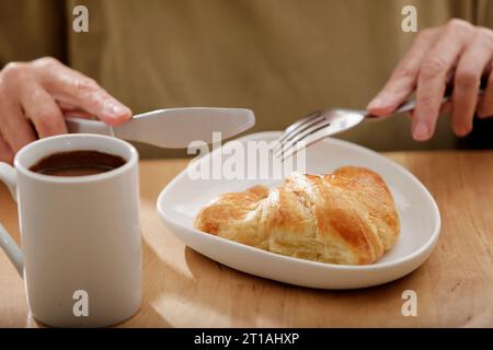 Frauenhände schneiden ein Croissant neben einer Kaffeetasse auf einer Teekanne aus hellem Holz Stockfoto