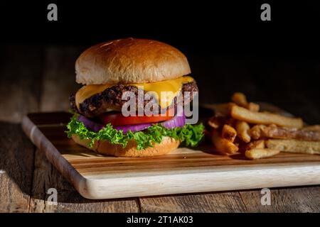 Ein frisch gegrillter Cheeseburger mit geschmolzenem Käse, Salat, Tomaten und Zwiebeln auf einem Brioche-Brötchen. Eine Seite Pommes Frites dazu Stockfoto