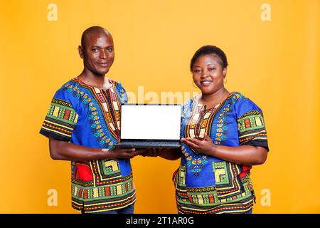 Lächelndes afroamerikanisches Paar, das ethnische Kleidung trägt und einen Laptop mit weißem leerem Bildschirm hält. Mann und Frau zeigen einen tragbaren Computer mit leerem Display und schauen in die Kamera Stockfoto