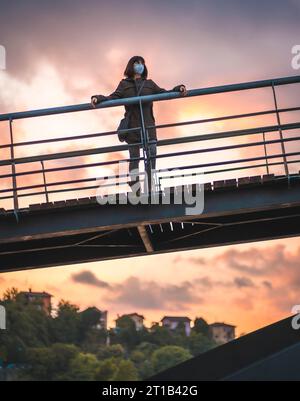 Eine junge brünette Frau mit einer Maske auf einer Brücke bei Sonnenuntergang. Erste Schritte der unkontrollierten COVID-19-Pandemie Stockfoto
