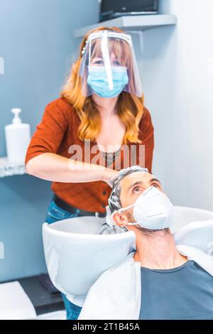 Friseure nach der Coronavirus-Pandemie. Kaukasischer Friseur mit Gesichtsmaske und Schutzschirm, covid-19. Soziale Distanz, neue Normalität. Stockfoto