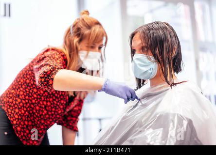 Friseur mit Maske und Handschuhen schneidet dem Kunden die Enden mit einer Schere ab. Wiederaufnahme mit Sicherheitsmaßnahmen für Friseure im COVID-19 Stockfoto