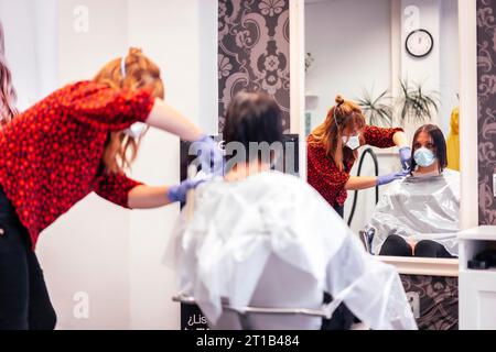Friseur mit Maske und Handschuhen schneidet die Enden zum Kunden, reflektiert im Spiegel. Wiederöffnung mit Sicherheitsmaßnahmen für Friseure im Stockfoto
