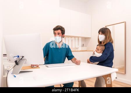 Arzt, der einen Computer benutzt, um den medizinischen Bericht einer Mutter mit Baby in den Armen in einem Krankenhaus zu zeigen Stockfoto