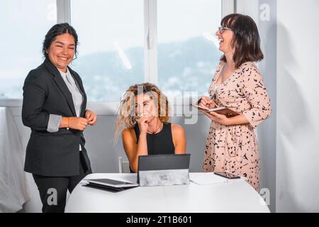 Junge Unternehmerin im Büro, zwei junge kaukasische Mädchen und eine junge Latina im Büro, die glücklich miteinander reden Stockfoto