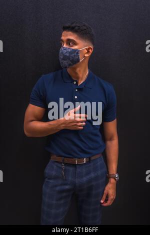 Mode-Lifestyle, Porträt eines jungen Latino-Mannes an einer schwarzen Wand. Blaues Poloshirt und karierte Hose. Bei einer Pandemie mit Maske Stockfoto