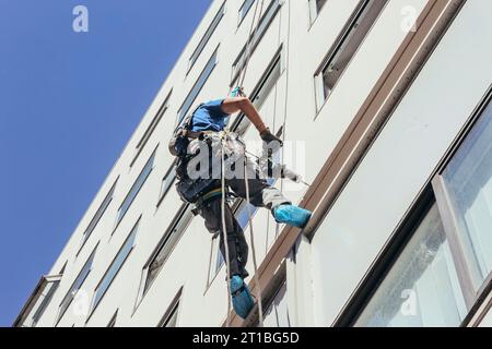 Arbeiter, der an einem Seil hängt und Lücken in der Fassade eines modernen Gebäudes schließt. Das Bergsteigerteam repariert die Fassade. Stockfoto