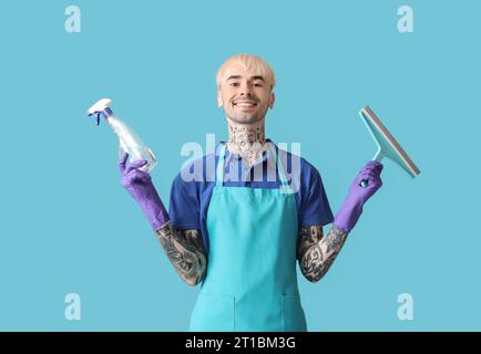 Junger tätowierter Mann mit Flasche Waschmittel und Rakel auf blauem Hintergrund Stockfoto