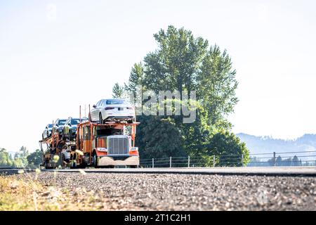 Orangefarbener Industrietransportwagen, großer Lkw mit verlängerter Fahrerkabine für Lkw-Fahrer, der Autos auf hydraulischem Auflieger transportiert, läuft Stockfoto