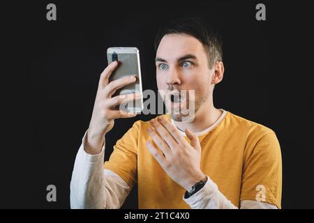 Schockierter junger kaukasischer Mann mit rosa schwarzem Hintergrund, offenem Mund, Telefonleseanzeige, unglaubliche große Einkaufspreise. Ein verblüffter Kerl liest das Stockfoto