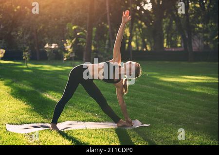 Die wunderschöne, sportlich geschnittene Yogi-Frau übt Yoga Asana Triangle Pose Trikonasana im Park bei Sonnenuntergang Stockfoto