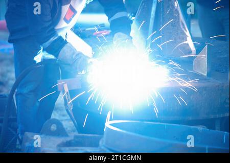 Industrieller Elektrodenschweißer mit Gesichtsschutz und blauem Gesamtschweißen eines Stahlrohrs in der Werkstatt. Nahansicht. Stockfoto