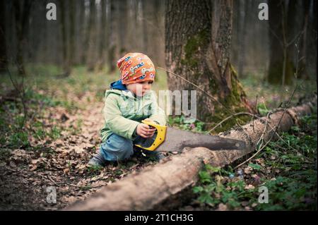 Kleiner Junge, der im Frühling im Wald einen umgestürzten Baum sägt Stockfoto