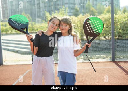 Kinder- und Sportkonzept. Porträt von lächelnden Mädchen, die auf dem Padel-Platz mit Schlägern und Tennisbällen im Freien posieren Stockfoto