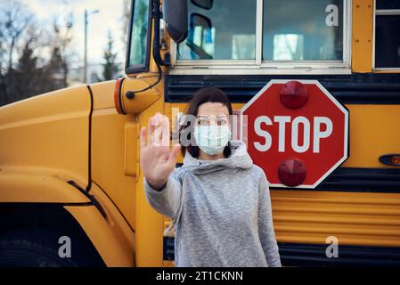 Eine Frau in Schutzmaske steht auf dem Hintergrund eines Schulbusses. Hebt die Hand und stoppt die Coronavirus-Epidemie. Ein großes Stoppschild ist visib Stockfoto