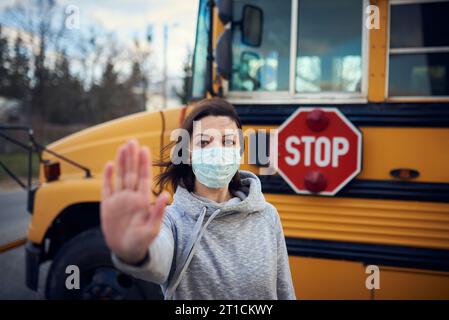 Eine Frau in Schutzmaske steht auf dem Hintergrund eines Schulbusses. Hebt die Hand und stoppt die Coronavirus-Epidemie. Ein großes Stoppschild ist visib Stockfoto