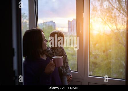 Mutter mit Baby schaut aus dem Fenster bei Sonnenuntergang in Isolation zu Hause für Virusausbruch. „Stay Home“-Konzept. Stockfoto