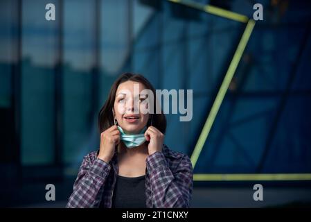 Junge glückliche Frau zieht schützende medizinische Maske draußen vor dem Hintergrund der modernen Stadt ab. Die COVID-19-Pandemie ist überwunden. Stockfoto