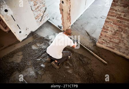 Männlicher Bauarbeiter, der Estrichschiene auf den mit Sand-Zement-Gemisch bedeckten Boden legt. Mann glättet und ebnet die Oberfläche mit dem Lineal beim Verputzen des Bodens in der Wohnung. Stockfoto