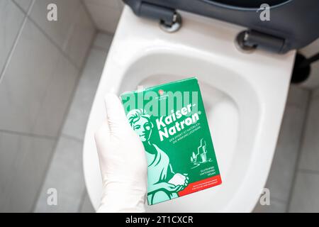 Augsburg, Bayern, Deutschland - 29. September 2023: Hand mit Handschuhen, die eine Packung Kaiser Natron vor einer Toilette hält. Hausmittel zum Reinigen und Entstopfen *** Hand mit Handschuhen hält eine Packung Kaiser Natron vor eine Toilette. Hausmittel zur Reinigung und zum lösen von Verstopfungen Stockfoto