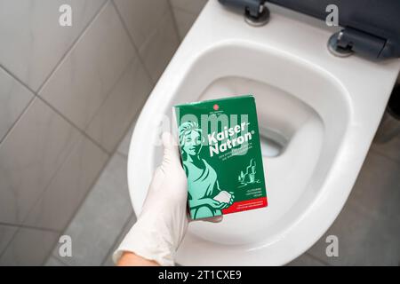 Augsburg, Bayern, Deutschland. September 2023. Handschuhe halten eine Packung Kaiser Natron vor einer Toilette. Hausmittel zum Reinigen und Entstopfen *** Hand mit Handschuhen hält eine Packung Kaiser Natron vor eine Toilette. Hausmittel zur Reinigung und zum lösen von Verstopfungen Credit: Imago/Alamy Live News Stockfoto