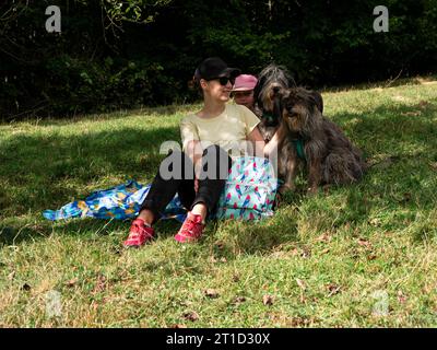 Glückliche Familienmutter mit Tochter und zwei Hunden, die auf grünem Rasen sitzen und ein lustiges Picknick machen, verbringen Zeit zusammen beim Wandern in den Bergen. Personen und Stockfoto