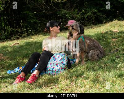 Glückliche Familienmutter mit Tochter und zwei Hunden, die auf grünem Rasen sitzen und ein lustiges Picknick machen, verbringen Zeit zusammen beim Wandern in den Bergen. Personen und Stockfoto