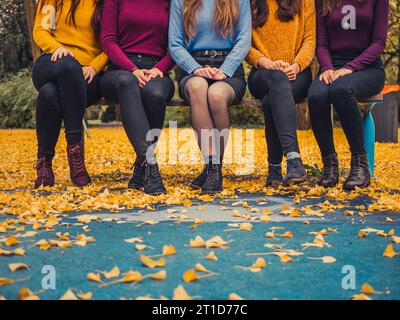 Fünf junge Freundinnen in lässiger modischer, trendiger, farbenfroher Kleidung Pullover Jeans sitzen auf einer Parkbank in herbstlichen bunten Blättern Stockfoto