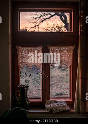 Altes ländliches Holzfenster mit Vorhängen von innen bei Sonnenuntergang Stockfoto