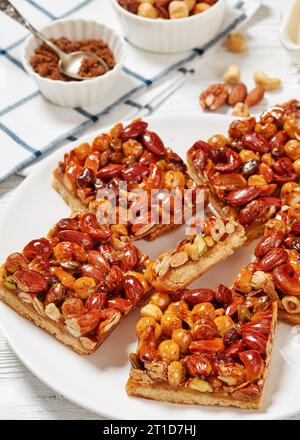 Hausgemachte Toffee gemischte Nüsse Shortbread Bars auf weißem Teller auf weißem Holztisch mit Zutaten, vertikale Ansicht, Nahaufnahme Stockfoto