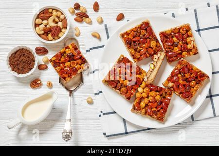 Hausgemachte Toffee gemischte Nüsse Shortbread-Bars auf weißem Teller auf weißem Holztisch mit Zutaten, horizontale Ansicht von oben, flache Lagen Stockfoto