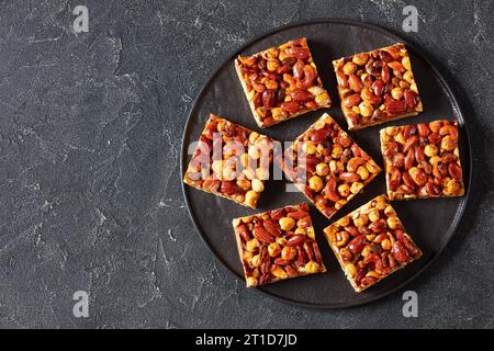 Hausgemachte Toffee gemischte Nüsse Shortbread Bars auf schwarzem Teller auf grauem Betontisch, horizontale Ansicht von oben, flache Lage, freier Platz Stockfoto