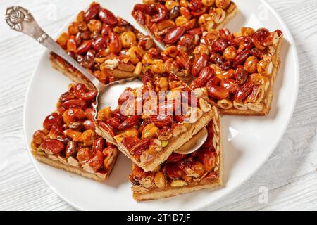 Altmodische Toffee gemischte Nüsse Shortbread Bars auf weißem Teller mit Kuchenschaufel auf weißem Holztisch, Nahaufnahme Stockfoto