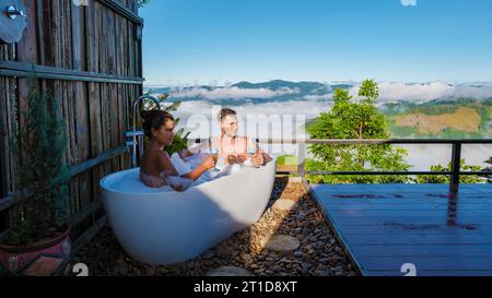 Ein multirassisches gemischtes Paar Männer und Frauen in einer Badewanne mit Blick auf die Berge von Chiang Rai Nord Thailand während des Urlaubs. Außenbadezimmer, Badewanne bei Sonnenuntergang Stockfoto