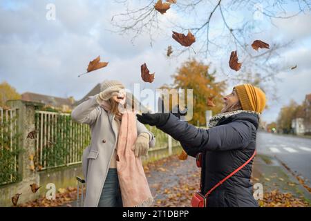 Ein paar glückliche Frauen, die in warmer Kleidung mit den Blättern in der Stadt spielen Stockfoto