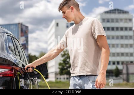 Mann, der eine Steckdose öffnet und ein Ladegerät in ein schwarzes Elektroauto an eine EV-Ladestation in der Stadtstraße stellt Stockfoto