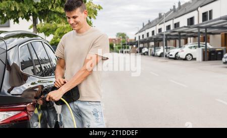 Mann, der eine Steckdose öffnet und ein Ladegerät in ein schwarzes Elektroauto an eine EV-Ladestation in der Stadtstraße stellt Stockfoto