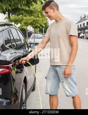 Mann, der eine Steckdose öffnet und ein Ladegerät in ein schwarzes Elektroauto an eine EV-Ladestation in der Stadtstraße stellt Stockfoto