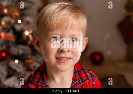 Der blonde Junge im roten karierten Pyjama schmückt Weihnachtskekse mit Glasur in der Küche. Stockfoto