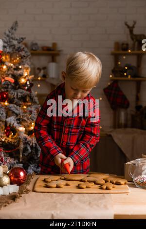 Der blonde Junge im roten karierten Pyjama schmückt Weihnachtskekse mit Glasur in der Küche. Stockfoto