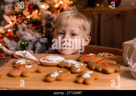 Der blonde Junge im roten karierten Pyjama schmückt Weihnachtskekse mit Glasur in der Küche. Stockfoto