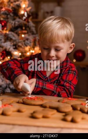 Der blonde Junge im roten karierten Pyjama schmückt Weihnachtskekse mit Glasur in der Küche. Stockfoto