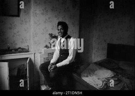 Der schwarze Mann saß auf seinem Bett in seinem viktorianischen Reihenhaus während der Slumräumung und des Abrisses von St Ann's, Nottingham, England. 1969-1970 Stockfoto