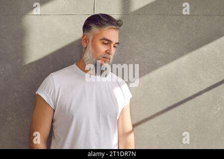 Hübsches älteres bärtiges männliches Modell in weißem T-Shirt, das an einer grauen Betonwand steht Stockfoto